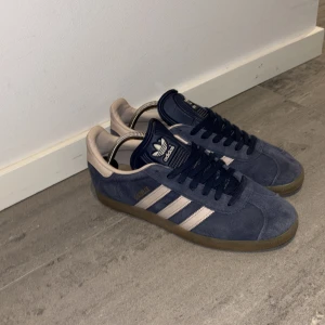 Blå Adidas Gazelle sneakers - Snygga Adidas Gazelle sneakers i blå mocka med beiga detaljer och klassiska tre ränder. Skorna har en gummisula och snörning för en bekväm passform. Perfekta för en stilren look. Skick 9/10 knappt använda.