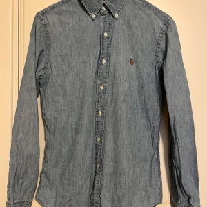 Blå jeansskjorta från Ralph Lauren - Snygg blå jeansskjorta från Ralph Lauren i slim fit. Skjortan har långa ärmar och knappar framtill samt en broderad logga på bröstet. Perfekt för en stilren look. Sparsamt använd i mycket gott skick. Se gärna mina andra annonser, så kan allt skickas i samma sändning. 