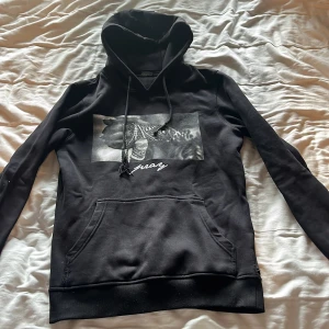 Svart hoodie från Mister Tee - Säljer en svart hoodie från Mister Tee med en cool tryckdetalj på framsidan. Hoodien har en känguruficka och justerbar huva med dragsko. Perfekt för en avslappnad stil.