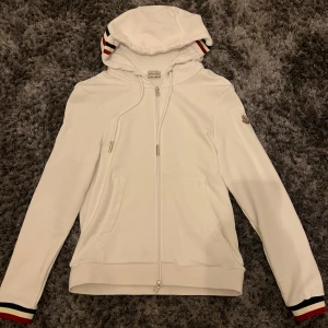 Vit huvtröja från Moncler - Säljer en stilren vit huvtröja från Moncler med dragkedja och randiga detaljer på ärmslut och huva. Perfekt för en casual look med en touch av lyx. Tröjan har långa ärmar och en bekväm passform.