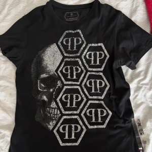 Svart t-shirt med dödskalle och logotyp - Snygg svart t-shirt från Philipp Plein med en glittrig dödskalle och logotypmönster på framsidan. Perfekt för dig som gillar en edgy stil. T-shirten har korta ärmar och en rund halsringning.  värt att nämna att några diamanter tappats från tröjan där av priset! (KAN GÅ NER I PRIS VID SNABB AFFÄR)