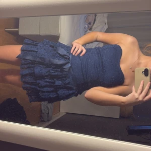 Mörkblå axelbandslös klänning - Snygg mörkblå axelbandslös klänning med volanger och spetsdetaljer. Klänningen har en dekorativ rosett i midjan och en kort längd som ger en festlig look. Perfekt för speciella tillfällen.