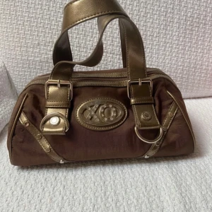 Brun och guld handväska - Snygg brun handväska med guldiga detaljer och handtag. Väskan har en dragkedja upptill och dekorativa spännen på sidorna. Perfekt för att ge en touch av glamour till din outfit.