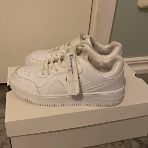 Vita sneakers från Champion - Snygga vita sneakers från Champion med snörning och kardborreband. Skorna har en klassisk design med Champion-loggan i blått och en skimrande detalj på sidan. Perfekta för en stilren look.