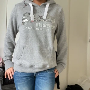 Grå hoodie med paljetter - Säljer en snygg grå hoodie från Superdry med paljettdetaljer på bröstet. Tröjan har en klassisk känguruficka och vita dragsnören i huvan. Perfekt för en avslappnad stil. Storlek S💗💗