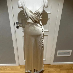 Fin långklänning Zara - Elegant champagnefärgad långklänning i satin. Klänningen har en v-ringad urringning och knappar längs sidan. Perfekt för speciella tillfällen.