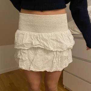 Vit kortkjol med volang - Säljer en söt vit kortkjol med volangdetaljer och elastisk midja. Kjolen har ett broderat mönster som ger en romantisk känsla. Perfekt för varma dagar och lätta att matcha med olika toppar.