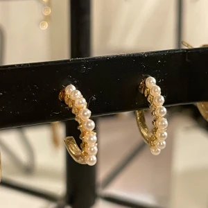 Guldörhängen med pärlor - Eleganta guldörhängen dekorerade med små vita pärlor. Perfekta för att ge en touch av lyx till din stil. De har en halvcirkelformad design som gör dem både moderna och tidlösa.