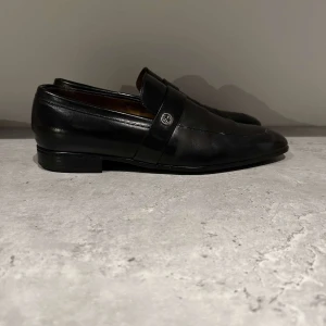 Svarta loafers från Gucci - Eleganta svarta loafers från Gucci i skinn med en diskret logotypdetalj på ovansidan. Skorna har en klassisk design med rundad tå och låg klack, vilket ger en stilren look. Perfekta för en sofistikerad stil.