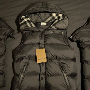 Burberry jacka - Säljer en snygg svart jacka från Burberry med avtagbara ärmar och en klassisk rutig huva. Perfekt för kyliga dagar! Tveka inte på att höra av er.