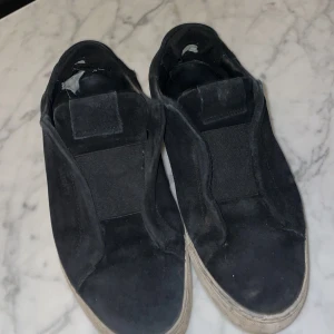 Svarta sneakers från Arigato - Säljer ett par svarta sneakers från Arigato i mocka. Skorna har en stilren design utan snörning och en vit sula. Perfekta för en avslappnad stil.