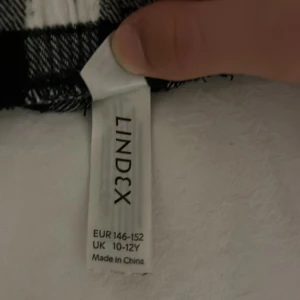 Svartvita rutiga byxor från Lindex - Säljer ett par mysiga svartvita rutiga byxor från Lindex i storlek 146-152. Perfekta för en avslappnad dag hemma eller som pyjamasbyxor. De har en elastisk midja med snörning för extra komfort. Materialet känns mjukt och skönt mot huden.