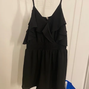 Svart playsuit med volangdetaljer - Snygg svart playsuit med volangdetaljer och smala axelband. Perfekt för festliga tillfällen. Den har en smickrande passform med elastisk midja som ger en fin siluett. 