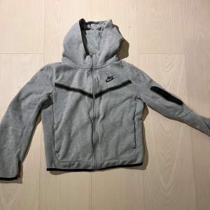 Säljer en grå hoodie från Nike i bra skick. Den har en dragkedja framtill och en huva för extra mysfaktor. Perfekt för höst och vår, med långa ärmar och en stilren design. Passar både till träning och vardag.