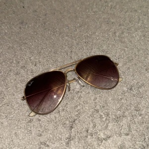 Ray-Ban Aviator Solglasögon - Snygga Ray-Ban aviator solglasögon med guldfärgad metallram och mörka tonade glas. Perfekta för en cool och klassisk look, oavsett om du är på stranden eller i stan. De har justerbara näskuddar för extra bekvämlighet.