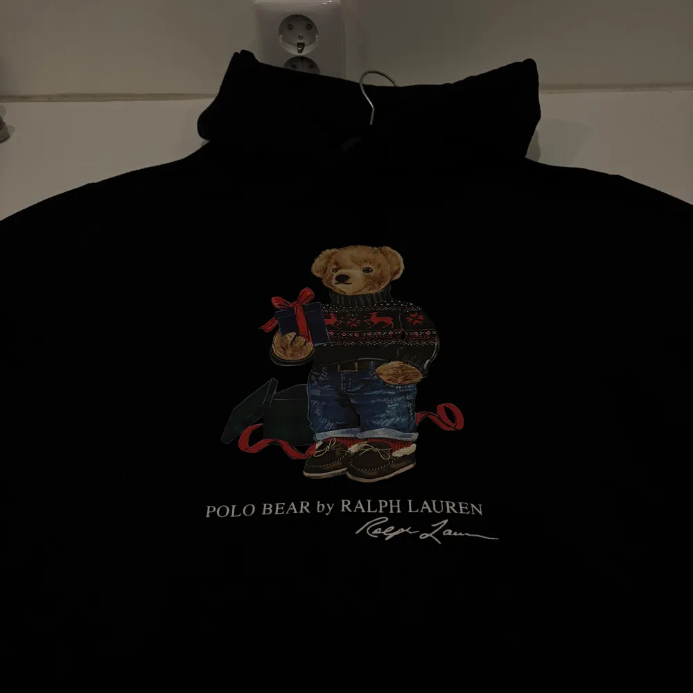 Säljer en svart hoodie från Ralph Lauren med den ikoniska Polo Bear på framsidan. Björnen har en jultröja och håller en present, vilket gör den perfekt för vintern och julen. Hoodien är i bra skick och har en mysig passform. Perfekt för att hålla sig varm och stilren under kalla dagar! Priset är ej hugget i sten, så vid smidig affär så går priset att sänkas👍🏻. Hoodies.