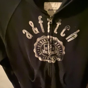 Svart hoodie från Abercrombie - Säljer en svart hoodie från Abercrombie med dragkedja och tryck på framsidan. Perfekt för en avslappnad stil och passar bra till både jeans och joggers. Den är i bra skick och redo för nya äventyr!