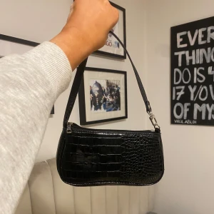 Svart axelväska med krokodilmönster - Snygg svart axelväska i krokodilmönstrat material. Väskan har en dragkedja upptill och en justerbar axelrem. Perfekt för en kväll ute eller en dag på stan. Den är både stilren och praktisk med gott om plats för dina nödvändigheter. Använd ett tal gånger, och därav priset! Dock så är den i perfekt material fortfarande- inga slitningar och ser helt ny ut. Köpt på bershka eller Urban outfitters för några år sen men inte använd idag.