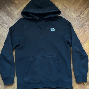 Stüssy Hoodie - Säljer en svart hoodie från Stüssy i storlek S. Den har en klassisk design med Stüssy-loggan tryckt både fram och bak. Pytte lite sprucket på märket, inget man märker vid användning. Den är även lite solblekt. Inga hål eller fläckar. Perfekt för höst och vinter.  Passar till allt, hör av dig vid frågor 👋