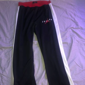 Svarta Jordan byxor med röd och vit detalj - Säljer ett par svarta Jordan byxor i mycket bra skick. De har en röd midja och vita ränder längs sidorna. Perfekta för träning eller en avslappnad stil. Jordan-loggan syns tydligt på sidan. Superbekväma och stilrena!