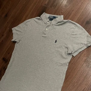 Grå pikétröja från Polo Ralph Lauren - Säljer en stilren grå pikétröja från Polo Ralph Lauren i custom fit. Tröjan har korta ärmar och en klassisk krage med knappar. Den är i mycket bra skick förutom den lilla skadan som syns på bild 4 och 5.