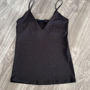Svart ärmlös linnetopp - Snygg svart linnetopp med smala axelband och v-ringning. Perfekt för sommardagar eller som ett basplagg i garderoben. Enkel och stilren design som passar till allt!