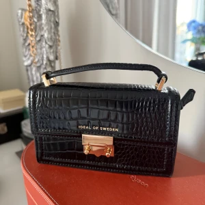 Svart handväska från Ideal of Sweden - Snygg svart handväska i krokodilpräglat mönster från Ideal of Sweden. Väskan har en elegant guldfärgad låsdetalj och ett praktiskt handtag. Perfekt för både vardag och fest!
