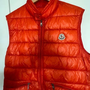 Orange dunväst från Moncler - Säljer en snygg orange dunväst från Moncler i mycket bra skick. Stoleken är 3 vilket motsvarar Medium. Västen har en dragkedja framtill och två praktiska fickor med dragkedjor. Perfekt för höst och vår när du vill hålla dig varm men ändå se stilren ut. Passar både till vardags och lite finare tillfällen.