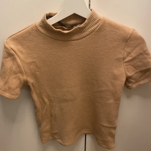 Beige kortärmad polotröja från Zara - Säljer en snygg beige kortärmad polotröja från Zara. Perfekt för både vardag och lite finare tillfällen. Den har en ribbad krage som ger en stilren look. Passar bra till jeans eller kjol för en chic outfit!