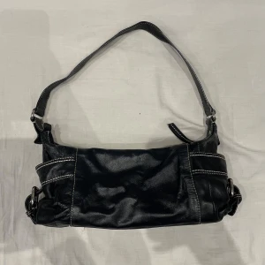 Svart axelväska i fejkskinn - Snygg svart axelväska i skinn med vita sömmar. Perfekt för vardagsbruk eller en utekväll. Väskan har en justerbar axelrem och stilrena detaljer. Den är i mycket bra skick och redo att användas direkt! Hittar inga uppenbara defekter 