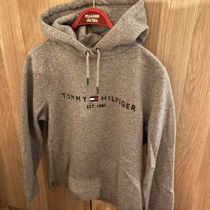 Säljer en snygg grå hoodie från Tommy Hilfiger med klassisk logga och text 'Est. 1985' på bröstet. Den har en bekväm passform med justerbar huva. Endast provad. I nyskick. 