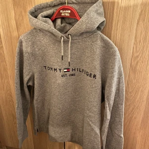 Tommy Hilfiger hoodie - Säljer en snygg grå hoodie från Tommy Hilfiger med klassisk logga och text 'Est. 1985' på bröstet. Den har en bekväm passform med justerbar huva. Endast provad. I nyskick. 