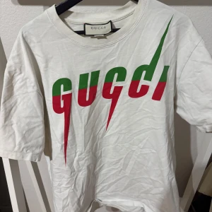 Vit t-shirt från Gucci - Säljer en snygg vit t-shirt från Gucci i storlek XL. Den har ett coolt tryck i grönt och rött på framsidan. Perfekt för sommaren eller våren. T-shirten är i bra skick och gjord av mjuk bomull. Passar perfekt till både vardag och fest!