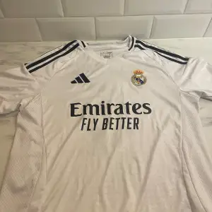 Säljer en vit Real Madrid fotbollströja i storlek L från Adidas. Tröjan har korta ärmar och är i mycket bra skick. Den har 'Bellingham' och nummer 5 tryckt på baksidan. Materialet är AEROREADY, vilket gör den perfekt för träning eller match. Perfekt för alla Real Madrid-fans! ⚽