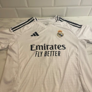 Vit Real Madrid fotbollströja från Adidas bytes:säljes - Säljer en vit Real Madrid fotbollströja i storlek L från Adidas. Tröjan har korta ärmar och är i mycket bra skick. Den har 'Bellingham' och nummer 5 tryckt på baksidan. Materialet är AEROREADY, vilket gör den perfekt för träning eller match. Perfekt för alla Real Madrid-fans! ⚽
