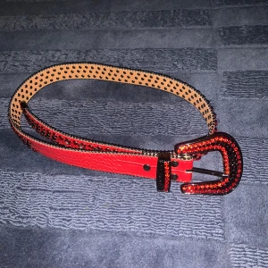 Rött bälte rhinestone  - Säljer ett coolt rött bälte med svarta nitar och glittrande strassdetaljer på spännet. Perfekt för att ge din outfit en edgy touch. Bältet är i mycket bra skick och passar perfekt till festliga tillfällen. Det har en unik design med både stil och attityd. Passa på att köpa detta statement-bälte!