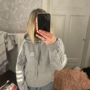 Grå hoodie från Adidas - Säljer en superbekväm grå hoodie från Adidas med klassiska vita ränder på ärmarna. Perfekt för chill dagar eller träning. Den har en stor ficka fram och en mysig huva. Passar perfekt till jeans eller träningsbyxor. 🩶
