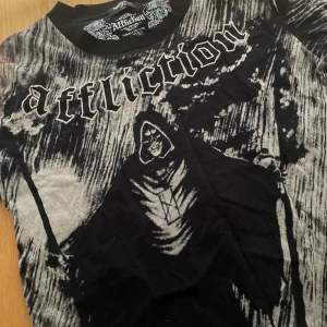 Affliction Tröja - Cool t-shirt från Affliction i svart och grått med ett mystiskt tryck av en figur i huva. Gjord i 100% bomull, perfekt för en avslappnad stil. Den har korta ärmar och en normal passform. Den har två fläckar på sidan men annars inget annat!  Perfekt för dig som gillar en edgy look! Köpte från en annan säljare på Plick! 💕