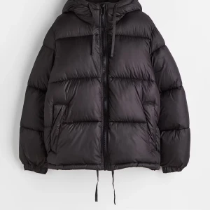 Svart pufferjacka med huva - Säljer en snygg svart pufferjacka med huva, perfekt för kalla dagar. Jackan har en dragkedja framtill och är långärmad. Den är i ett puffigt material som håller dig varm och bekväm. Passar perfekt! Skriv för fler bilder