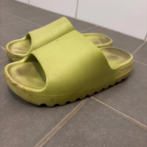 Gröna tofflor liknande till yeezy slides, strl 38 - Säljer ett par gröna sandaler i syntetmaterial och är i strl 38. De har en enkel och stilren design med öppen tå och en bred rem över foten. Perfekta för sommardagar eller att glida runt hemma. De är i bra skick men har några tecken på användning. Sköna och lätta att bära!