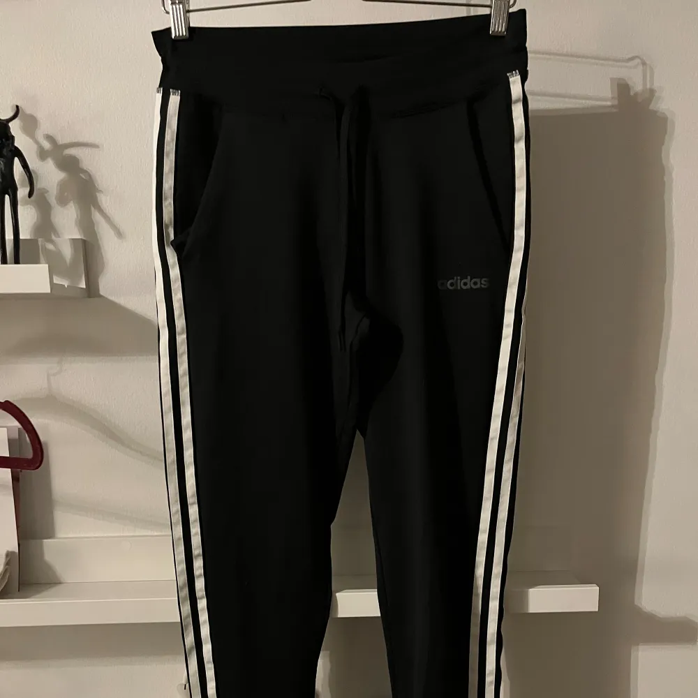 Säljer ett par svarta träningsbyxor från Adidas i storlek XS. Byxorna har vita ränder längs sidorna och en bekväm dragsko i midjan. Perfekta för träning eller en avslappnad dag. De är i mycket bra skick. . Sportswear.