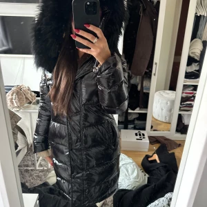Svart glansig dunjacka med päls - Säljer en superfin svart glansig dunjacka med stor fluffig päls på huvan. Perfekt för kalla vinterdagar! Jackan har en dragkedja framtill och långa ärmar. Den är både snygg och varm, perfekt för vinterns alla äventyr. Passar lika bra till vardags som till festliga tillfällen.