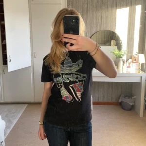 Svart t-shirt från Adidas - Säljer en svart t-shirt från Adidas i bra skick. Den har ett coolt tryck med färgglada sneakers och Adidas-loggan på framsidan. Bra passform och tyget är ganska tjockt
