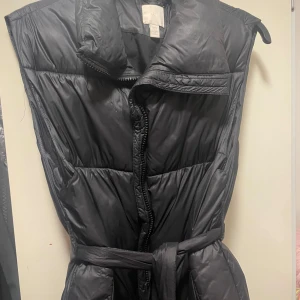 Svart dunväst med bälte - Säljer en svart dunväst i mycket bra skick. Västen har en dragkedja framtill och ett bälte i midjan för en snygg passform. Perfekt för höst och vinter när du vill hålla dig varm och stilren. Passar bra till både vardag och fest!