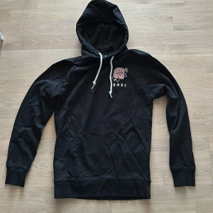 Svart hoodie med rosdetalj - Säljer en svart hoodie från Dope med tryck på bröstet och ryggen. Strlk XS