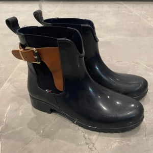 Stövlar från Tommy Hilfiger - Snygga marinblå stövlar från Tommy Hilfiger i syntetmaterial med bruna detaljer och ett dekorativt bälte. Perfekta för höst och vår med en robust sula för bra grepp. De har en stilren design som passar både till vardags och lite finare tillfällen. Storlek 37, normal i storleken.