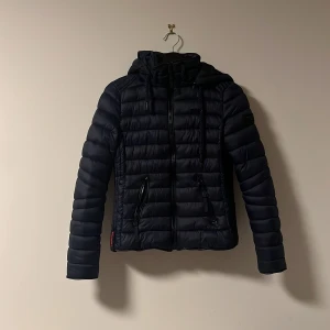 Svart pufferjacka från Navahoo - Säljer en svart pufferjacka från Navahoo i mycket bra skick. Jackan har en snygg quiltad design med dragkedja och huva, perfekt för kyliga höst- och vinterdagar. Den är långärmad och har en normal passform. Supermysig och stilren för alla tillfällen! Bara testad förra vintern och sen bara hängt i garderoben för den va för liten. 