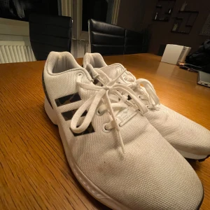 Vita sneakers från Adidas - Säljer ett par vita Adidas sneakers i bra skick. De har några detaljer och klassisk snörning. Perfekta för vardagsbruk och träning. Sulan är robust och ger bra grepp. Passar till både jeans och träningskläder.