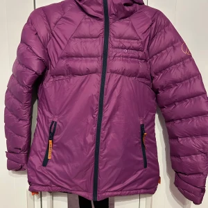 Lila dunjacka från Stormberg - Säljer en snygg lila dunjacka från Stormberg i mycket bra skick. Jackan har en dragkedja framtill och praktiska fickor med dragkedjor. Perfekt för kalla vinterdagar med sin varma och sköna passform. Den har en cool logga på ärmen och en justerbar huva. Passar perfekt för både vardag och utomhusaktiviteter.