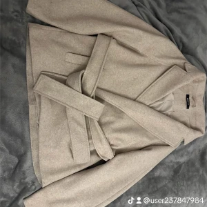 Beige kappa med bälte - Säljer en stilren beige kappa i mycket bra skick. Den har ett bälte i midjan för en snygg passform och långa ärmar. Perfekt för höst och vår när du vill hålla dig varm och chic. Passar både till vardags och festliga tillfällen. Storlek M, använd lite men syns knappt